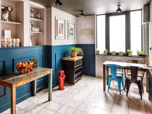 A dining area with rustic wooden tables, dark blue panelled walls, a red fire hydrant, a tiled floor and tall windows letting in daylight.