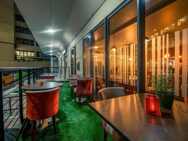 A covered outdoor terrace at night with green artificial turf, round tables and red upholstered chairs beside floor-to-ceiling glass windows.