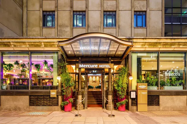 The front entrance of the Mercure Hotel at dusk, with a glass canopy, potted plants and illuminated shopfront windows.