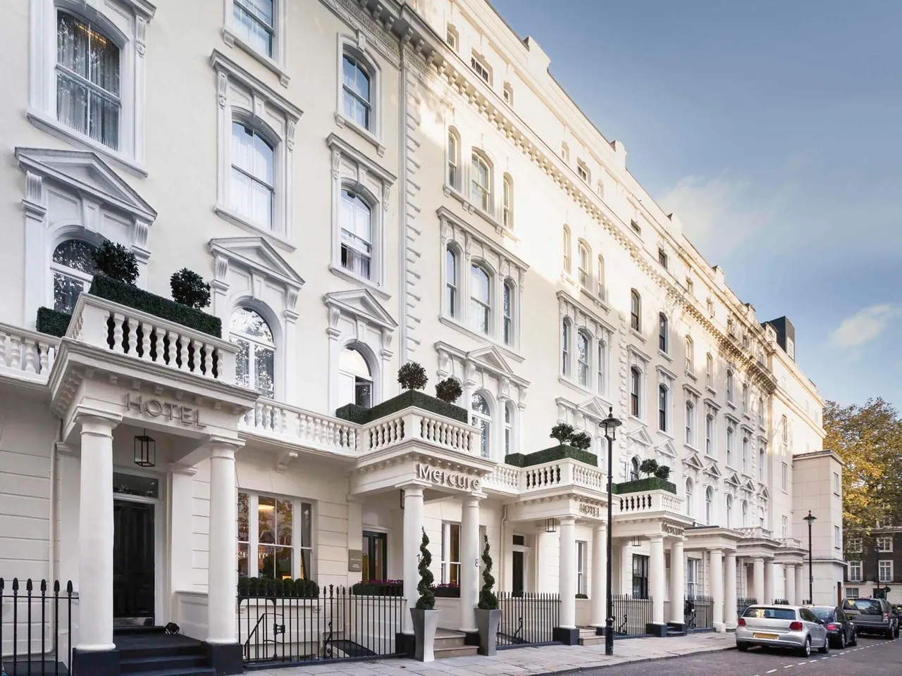 A white stucco terraced hotel building with Hotel and Mercure signage, black railings, a street lamp and parked cars along the road.
