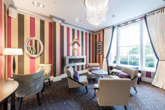 A lounge with striped wallpaper, a crystal chandelier, upholstered armchairs and a tall window looking onto trees and hedges.