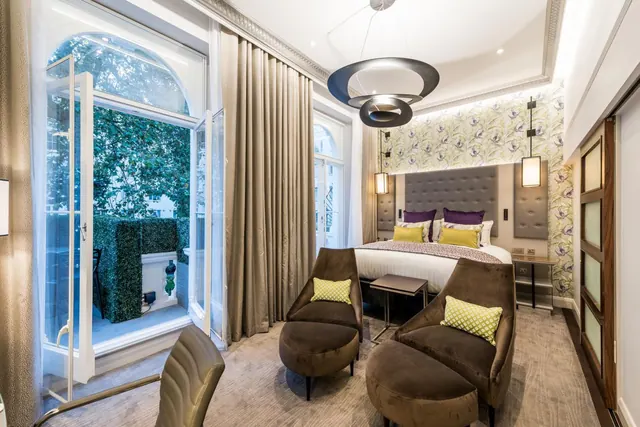 A hotel bedroom with a double bed, two armchairs with ottomans, and tall arched windows looking out onto trees and a hedge.