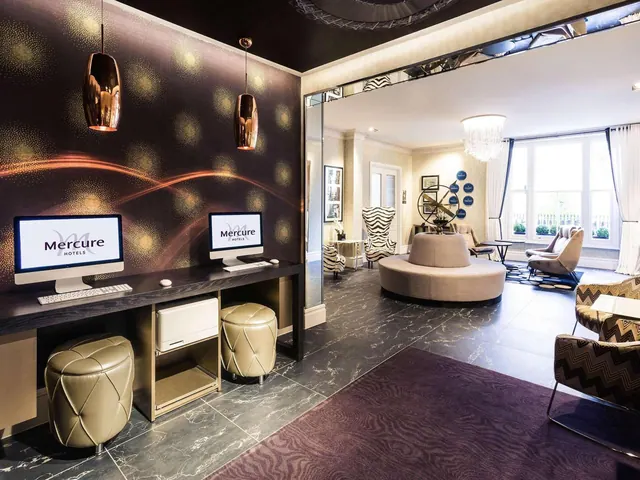 A hotel lobby shows a computer workstation with two monitors against a dark patterned wall, with a round sofa, zebra-print armchairs and a chandelier in the seating area beyond.