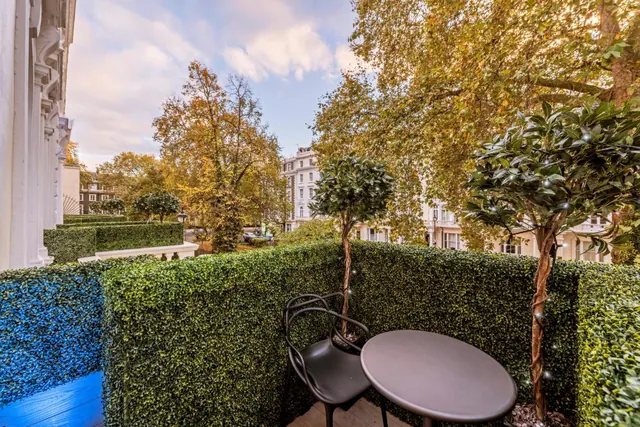 A small private balcony with hedging, a round table and chair, and topiary trees, looking out onto a tree-lined street with terraced buildings.