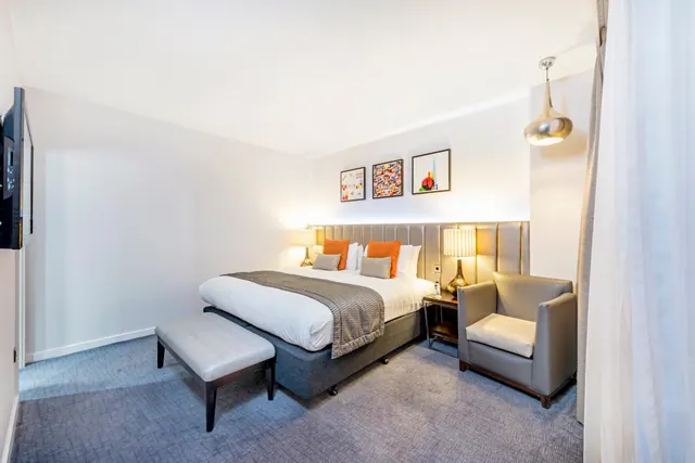 A bedroom with a made double bed with grey and orange cushions, a padded bench at the foot of the bed, a grey armchair, bedside lamps and a wall-mounted television.
