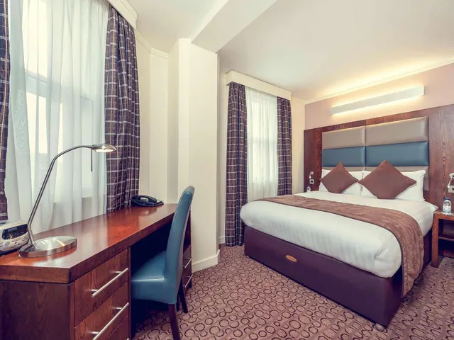 A double bed with brown cushions and a throw sits across from a wooden desk, blue chair and desk lamp in a hotel bedroom.