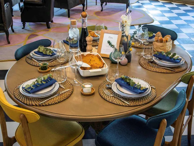 A set dining table with blue napkins, wine glasses, a casserole dish and a bread basket, with a checkered floor and armchairs in the background.