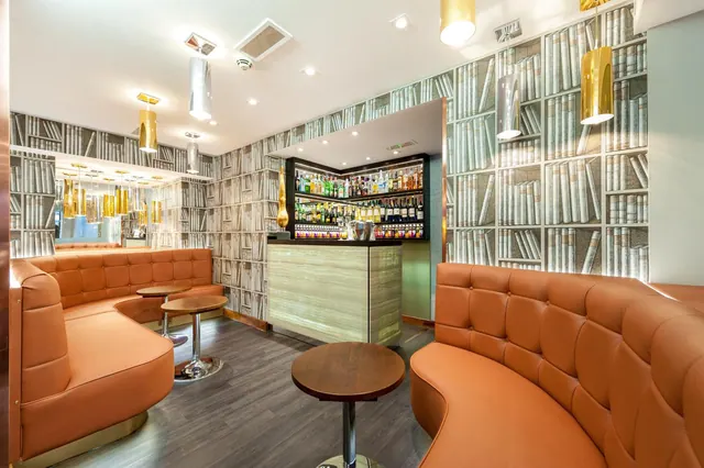 A bar lounge with curved tufted orange leather banquettes, round wooden tables, a bottle-lined bar counter and bookshelf-patterned wallpaper.