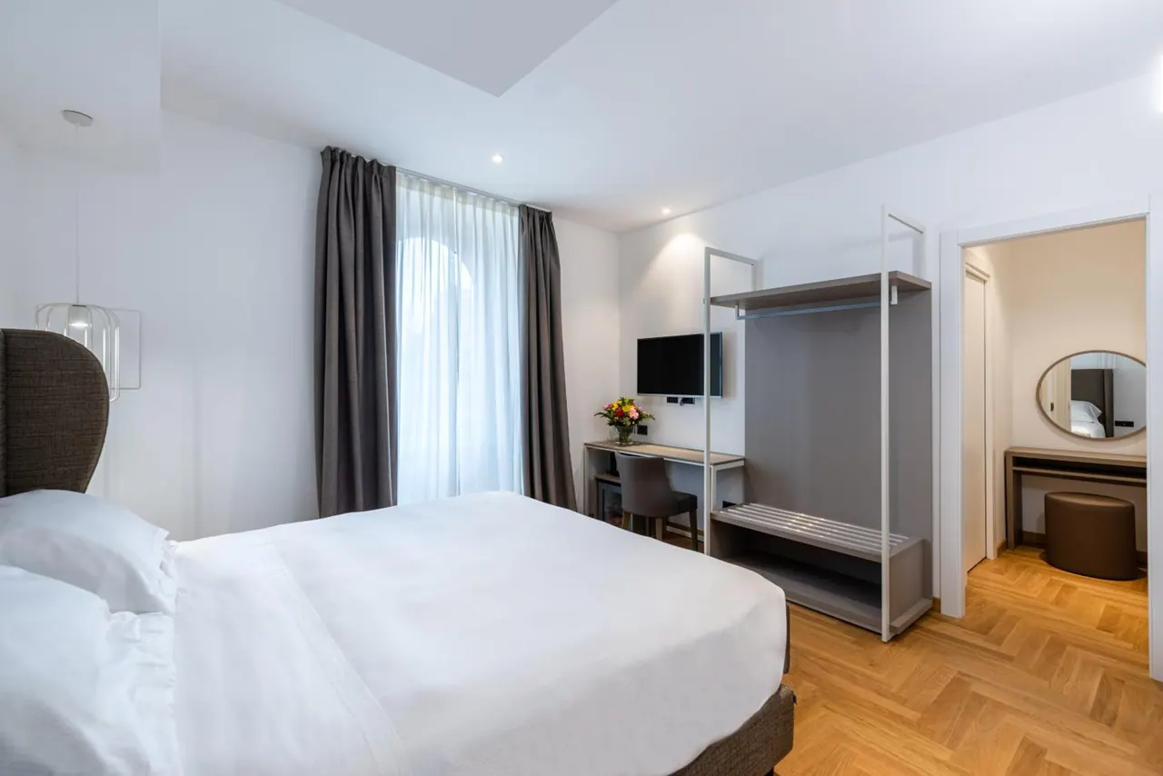 A bedroom with a large white bed, a wall-mounted television, a desk with a chair, an open wardrobe rail, and a doorway to a vanity area with a round mirror.