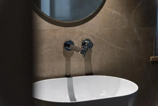 A wall-mounted chrome tap and an oval white basin sit below a round mirror, against beige stone-look tiles in a bathroom.