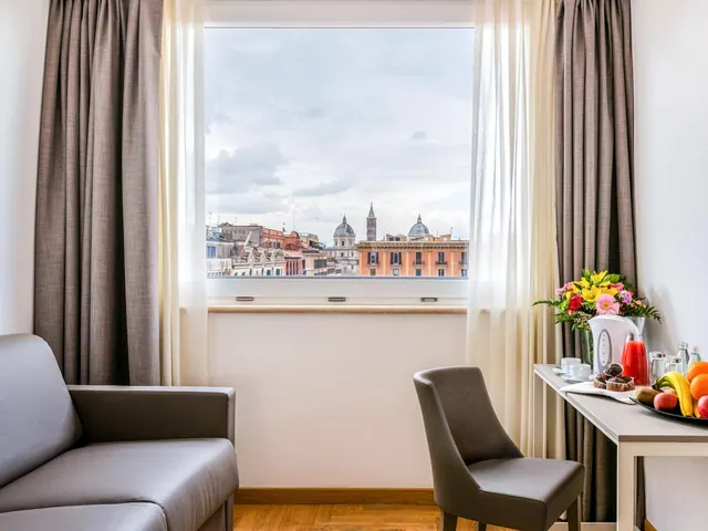 A hotel room with a grey sofa and a desk holding fruit, flowers and juice, with a window showing Rome rooftops and domes.