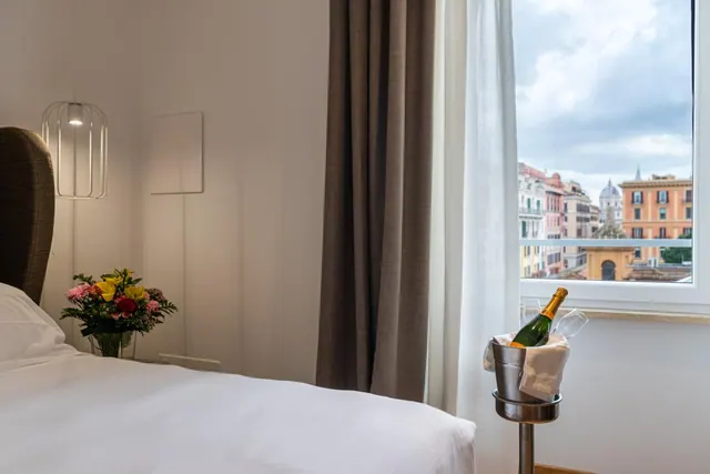 A made bed with white linen sits beside a window looking out over Rome's rooftops and a domed church, with a flower arrangement and a champagne bottle in an ice bucket on a stand.