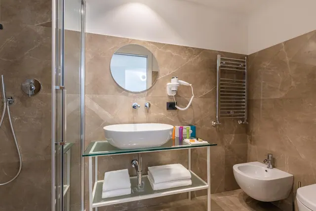 A bathroom with a glass-enclosed walk-in shower, a vessel sink on a glass shelf with towels underneath, a wall-mounted bidet and a heated towel rail.