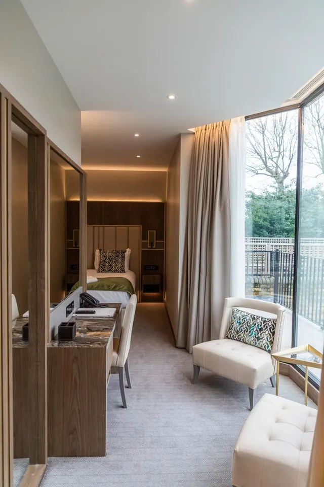 A narrow hotel bedroom with a single bed at the far end, a desk beside mirrored wardrobe doors, and white upholstered chairs by a floor-to-ceiling window looking onto trees and a railing.