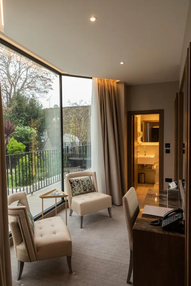 A hotel room with two upholstered armchairs by floor-to-ceiling windows looking onto trees and a garden, with a desk and an open bathroom doorway on the right.