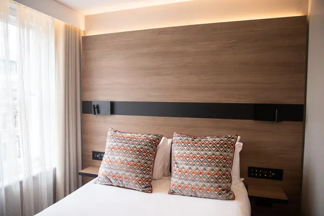 A double bed with two patterned cushions sits in front of a wood-panelled headboard wall, with a sheer-curtained window to the left.