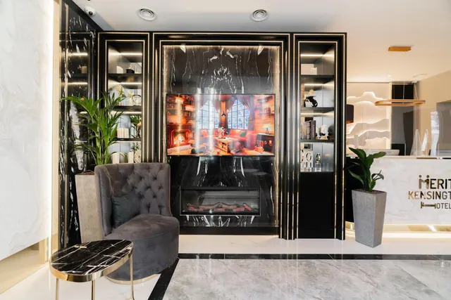 A hotel lobby with a grey tufted armchair, a wall-mounted television above an electric fireplace, and a reception desk with partial signage reading Merit Kensington Hotel.