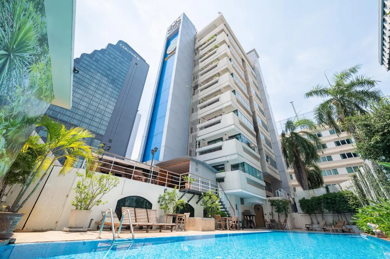 A pool with sun loungers and a pool ladder sits in front of a tall hotel tower with balconies, with a palm tree and a neighbouring office tower behind it.