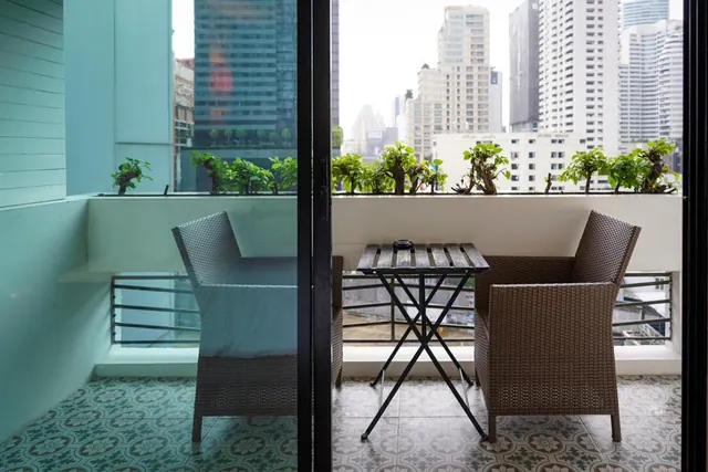 A balcony with two wicker chairs, a small folding table and potted shrubs overlooks high-rise towers under an overcast sky.