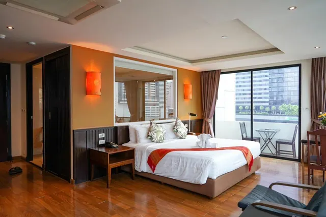 A bedroom with a king bed, an orange accent wall, and a sliding door opening onto a balcony with chairs and a view of high-rise buildings.