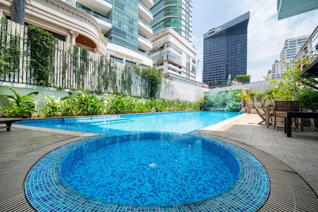 A large rectangular pool and a round mosaic-tiled pool sit on a terrace, with tall buildings and plants behind them.