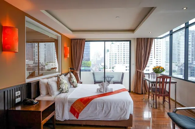 A bedroom with a made king bed, a work desk and armchair by floor-to-ceiling windows, and high-rise buildings visible beyond a sliding door to a balcony.