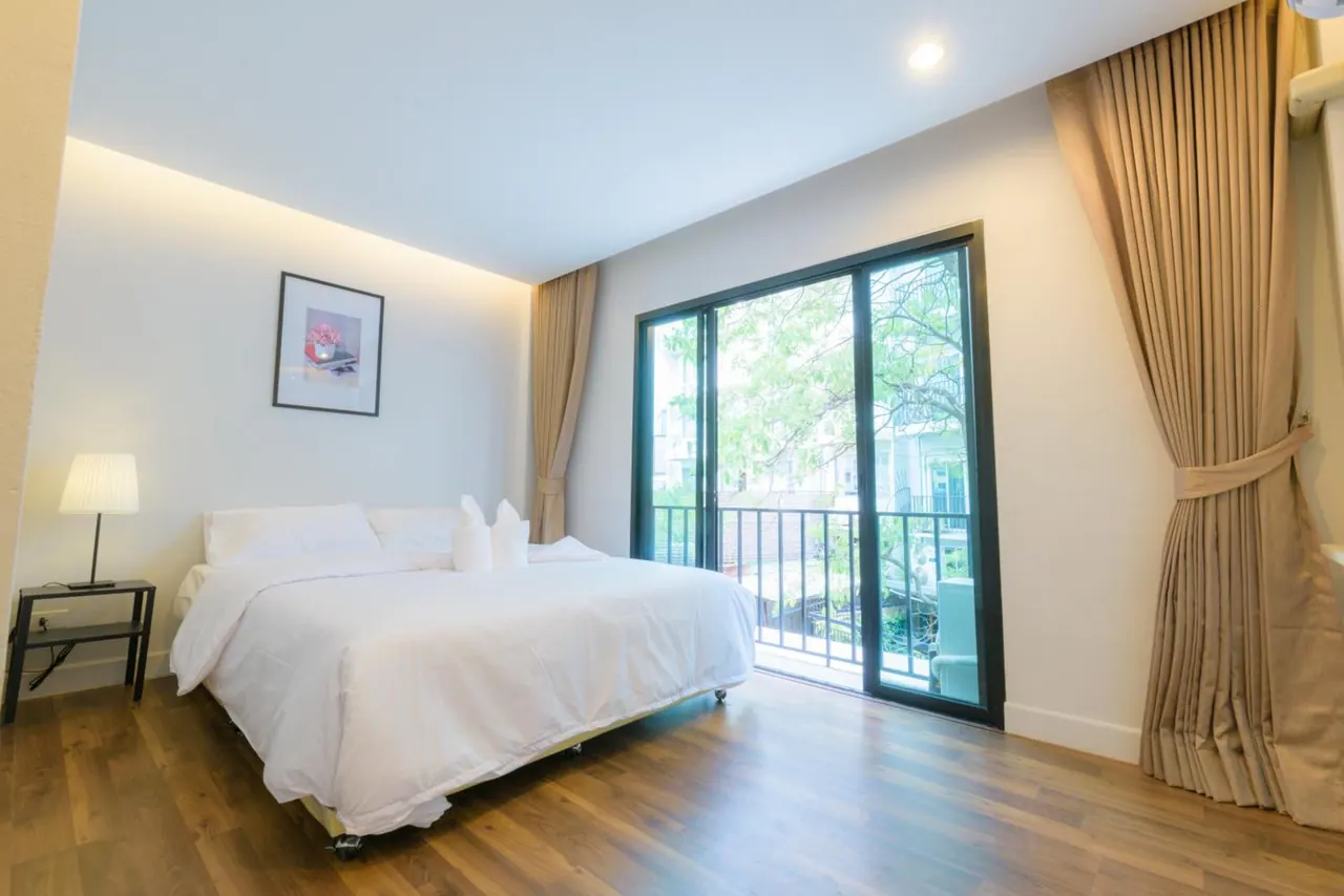 A bedroom with a made white double bed, a side table with a lamp, and sliding glass doors opening onto a balcony with trees beyond.