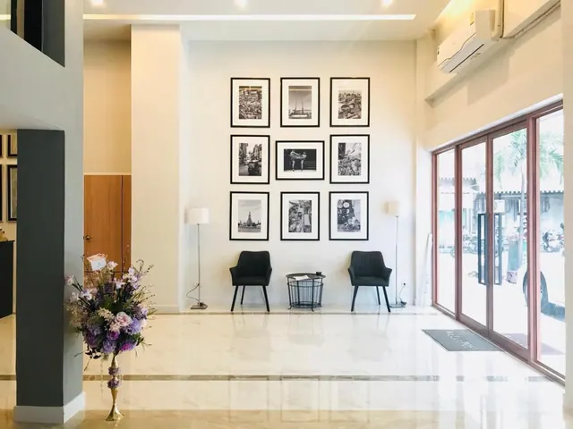 A lobby with a gallery wall of framed black-and-white photographs, two armchairs with a side table, a tall flower arrangement, and glass doors opening onto an outdoor parking area.