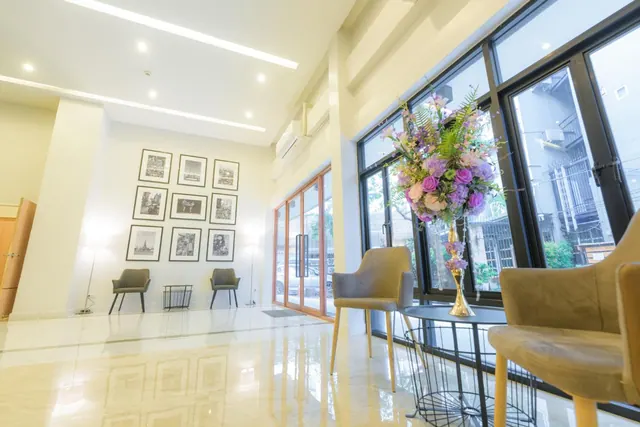 A lobby with a polished marble floor, framed black and white photographs on a white wall, armchairs, a floral arrangement on a black table, and tall windows onto a neighbouring building.