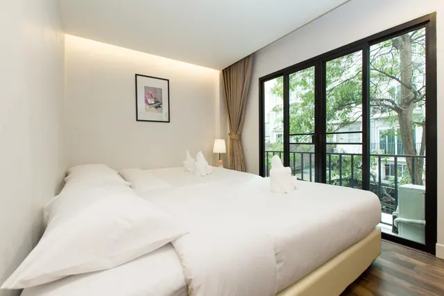 A bedroom with a large white bed topped with two towel swans, and black-framed glass doors opening onto a balcony with trees and a neighbouring building outside.