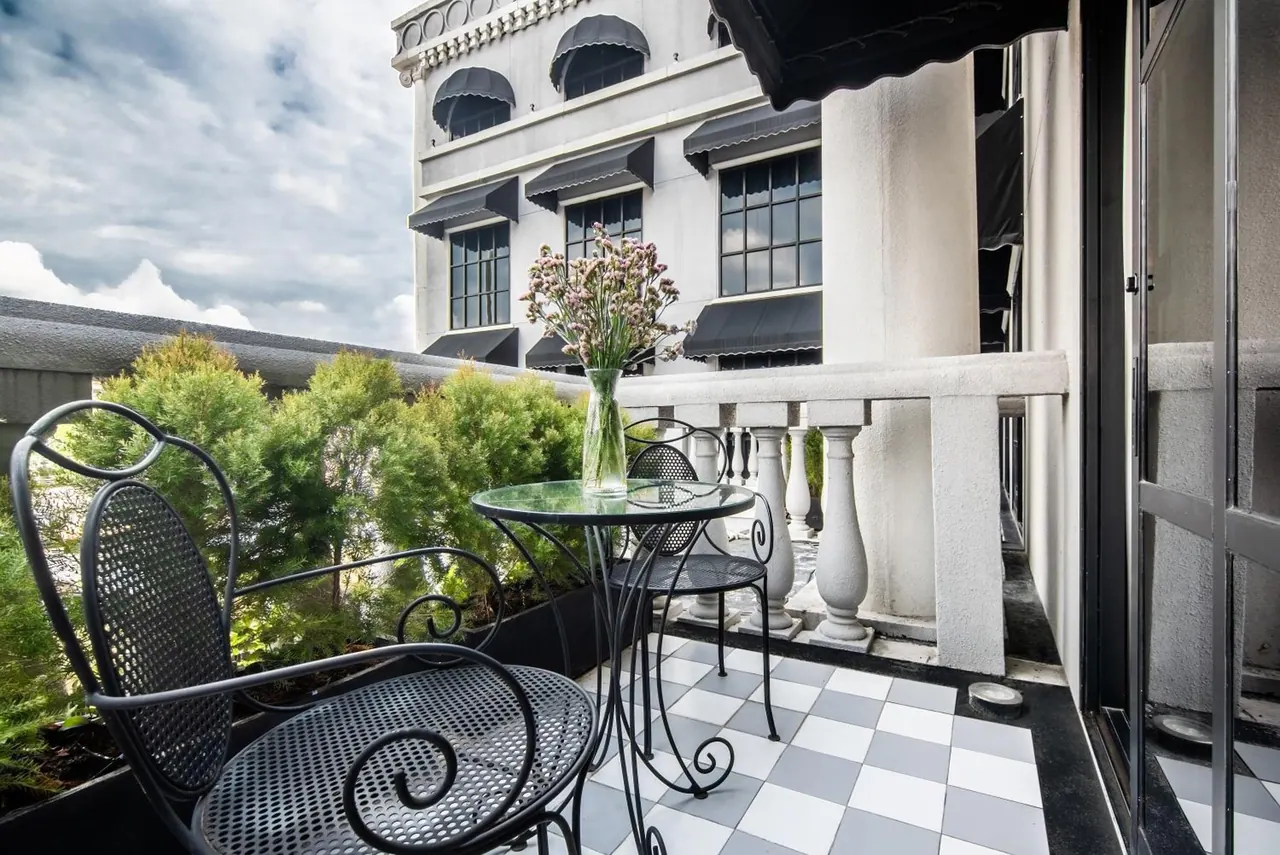 A balcony with a checkered tile floor, a wrought iron bistro table and two chairs, a vase of flowers, and a white balustrade, with a classical building facade and potted greenery beyond.