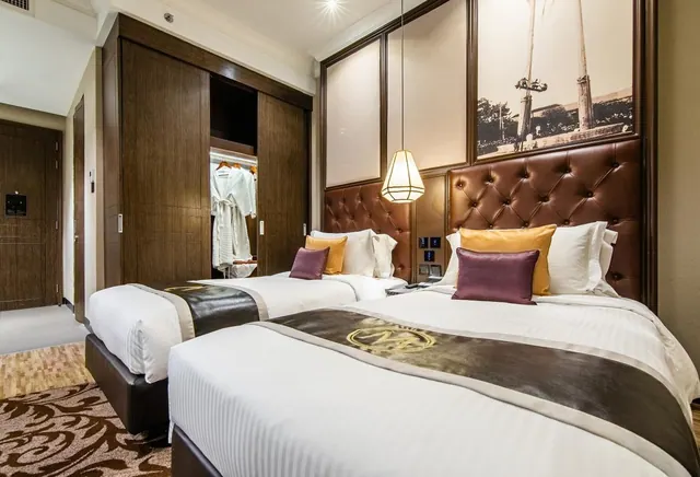 A hotel bedroom with two twin beds under a tufted brown leather headboard, a framed sepia photograph on the wall, and an open wardrobe holding white robes.