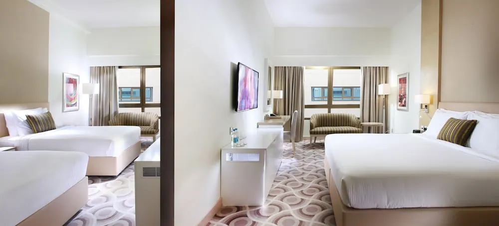 A hotel bedroom with two twin beds on the left and a king bed on the right, a wall-mounted television, armchairs by the windows, and a white desk.