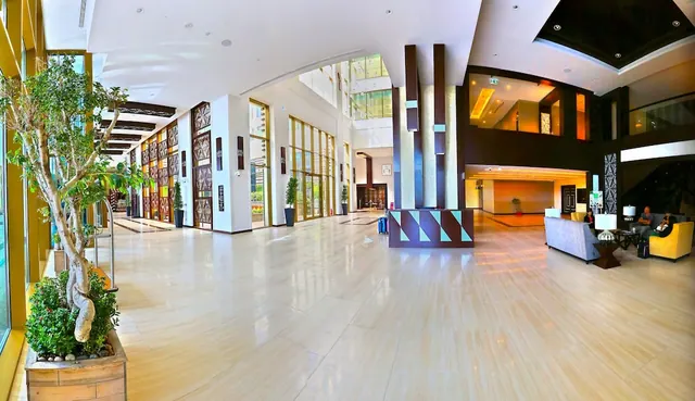 A wide-angle view of a hotel lobby with a polished stone floor, a seating area with sofas and armchairs on the right, a potted tree on the left, and two people seated.