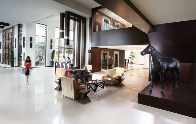 A hotel lobby with a seating area of armchairs and low tables, a black horse sculpture on a raised platform, and a lift bank beside it.