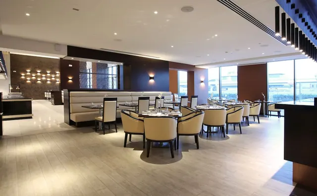 A restaurant dining room with cream leather armchairs, a built-in banquette along the back wall, set tables and floor-to-ceiling windows on the right.