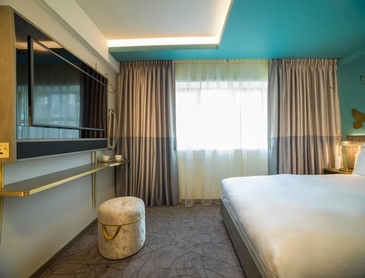 A bedroom with a large white bed, a wall-mounted television, a cushioned pouf and a window with sheer curtains.