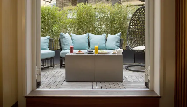 View through open French doors onto a private terrace with a blue-cushioned outdoor sofa, a coffee table holding drinks and plates, a hanging wicker egg chair, and a bamboo screen behind.