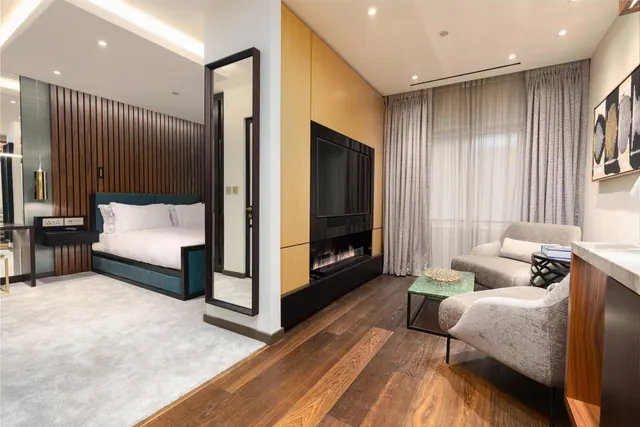 A hotel suite with a made bed visible on the left and a living area on the right, with a wall-mounted television above an electric fireplace and two armchairs beside sheer curtained windows.
