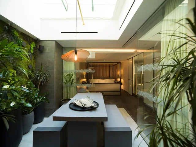 A concrete dining table with a bench and a black plate holding a decorative leaf sits under a skylight and pendant lamp, with a bed visible through glass partitions in the background.