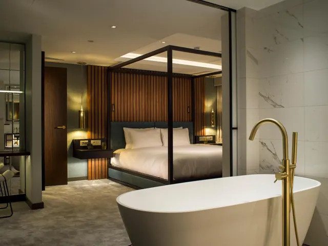 A freestanding bath with a brass floor-mounted tap sits in the foreground, with a canopy bed with white linen and a wooden slatted wall in the room beyond.