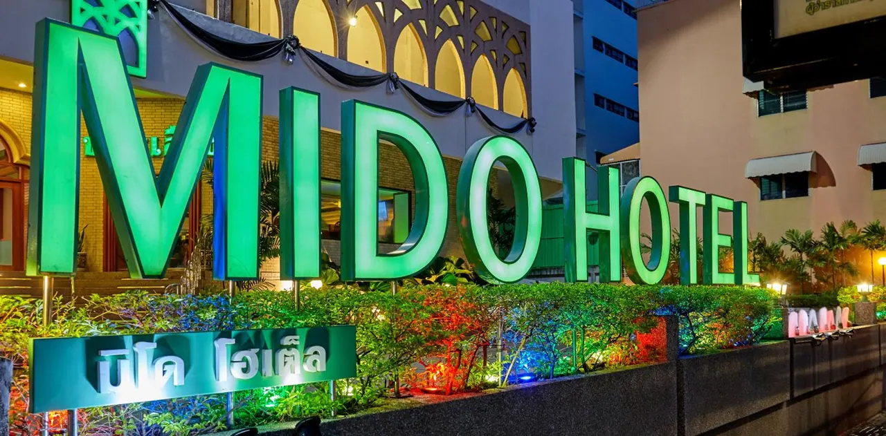 The illuminated green MIDO HOTEL sign stands above a planted hedge at the front of the building at night.