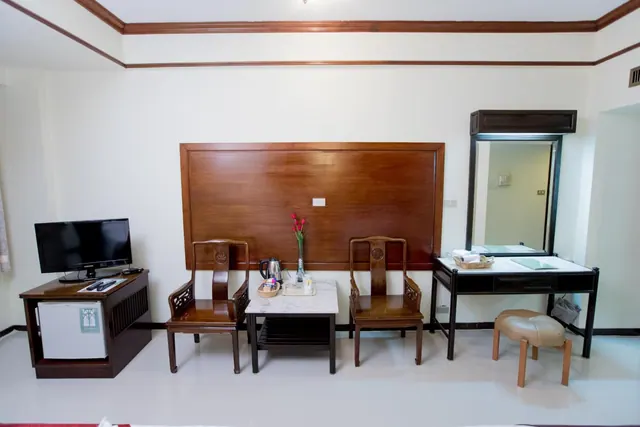 A hotel room with a wooden accent wall, a flat-screen television on a wooden cabinet above a mini fridge, two carved wooden armchairs, a marble side table with an electric kettle, and a dressing table with a mirror.