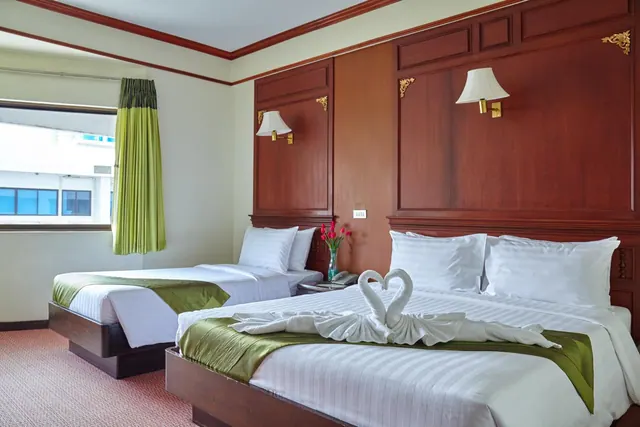 A hotel bedroom with a double bed dressed with towel swans, a single bed beside it, a wood-panelled headboard wall, and a window with a green curtain looking onto a neighbouring building.