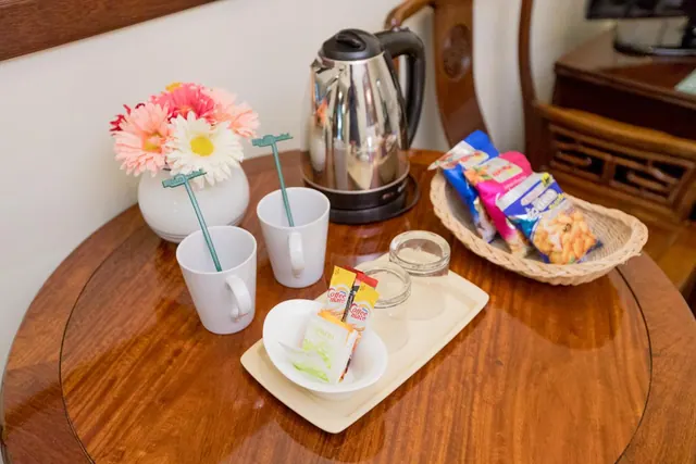 A round wooden table holds an electric kettle, two white mugs with stirrers, a flower vase, sugar and creamer sachets, glass tumblers, and a wicker basket of snack packets.