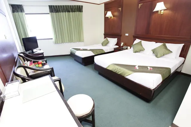 A hotel bedroom with a double bed and a single bed, a flat-screen TV on a desk, armchairs, a fruit plate on a table, and green curtains by the window.