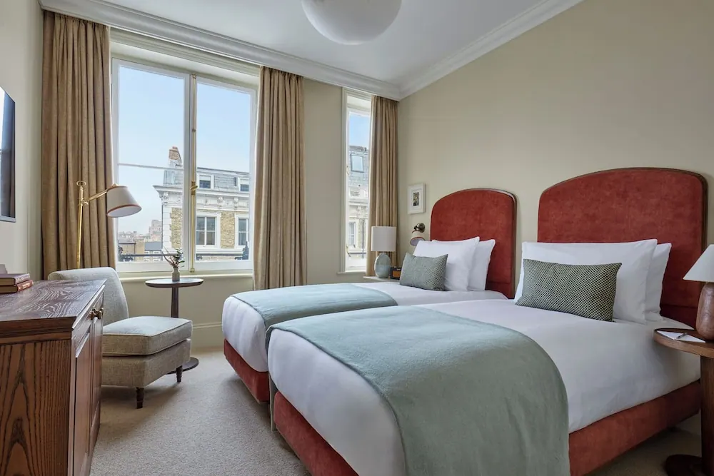 A twin bedroom with two beds with red upholstered headboards, an armchair, a wooden cabinet and tall windows looking onto neighbouring period buildings.