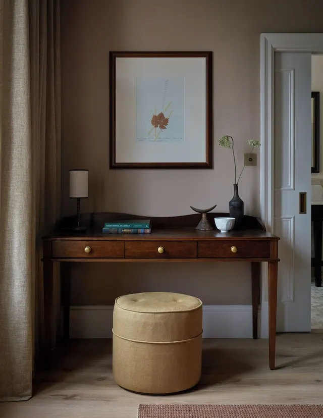 A wooden desk with three drawers stands against a wall beneath a framed botanical print, with a table lamp, vase, bowl and upholstered pouf nearby.
