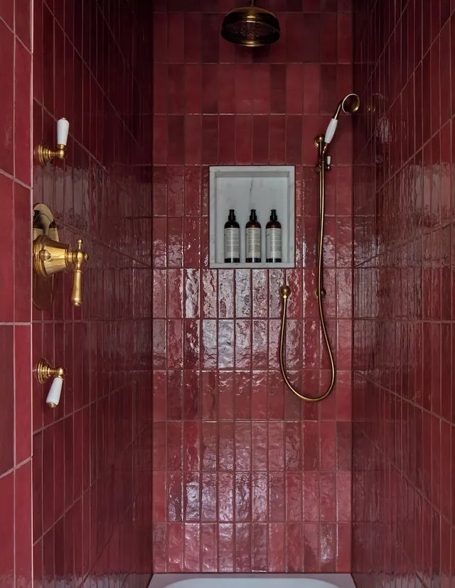 A walk-in shower with glossy red wall tiles, brass fittings, a overhead rain head and a handheld shower, and a recessed niche holding three toiletry bottles.