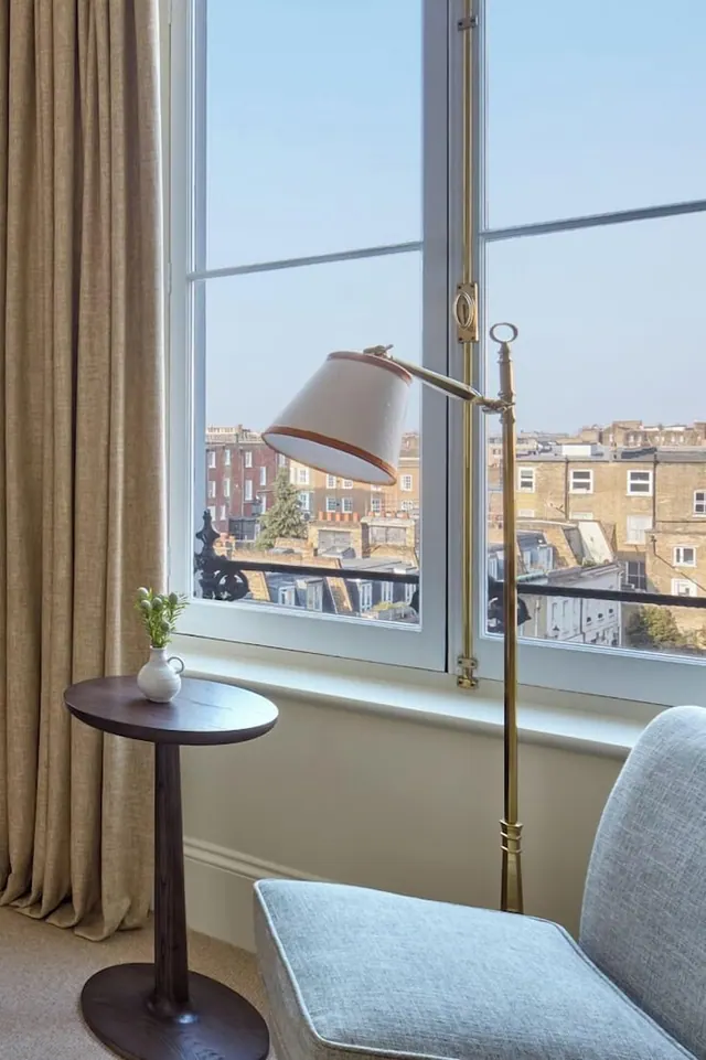 A seating corner beside a sash window with a brass floor lamp and a small wooden side table holding a vase, with a view over London rooftops and brick townhouses.